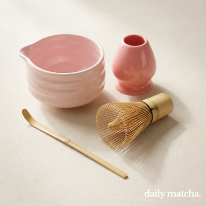 Pink ceramic bowl, small pink container, gold whisk, and wooden scoop on a light surface with 'daily matcha' branding.